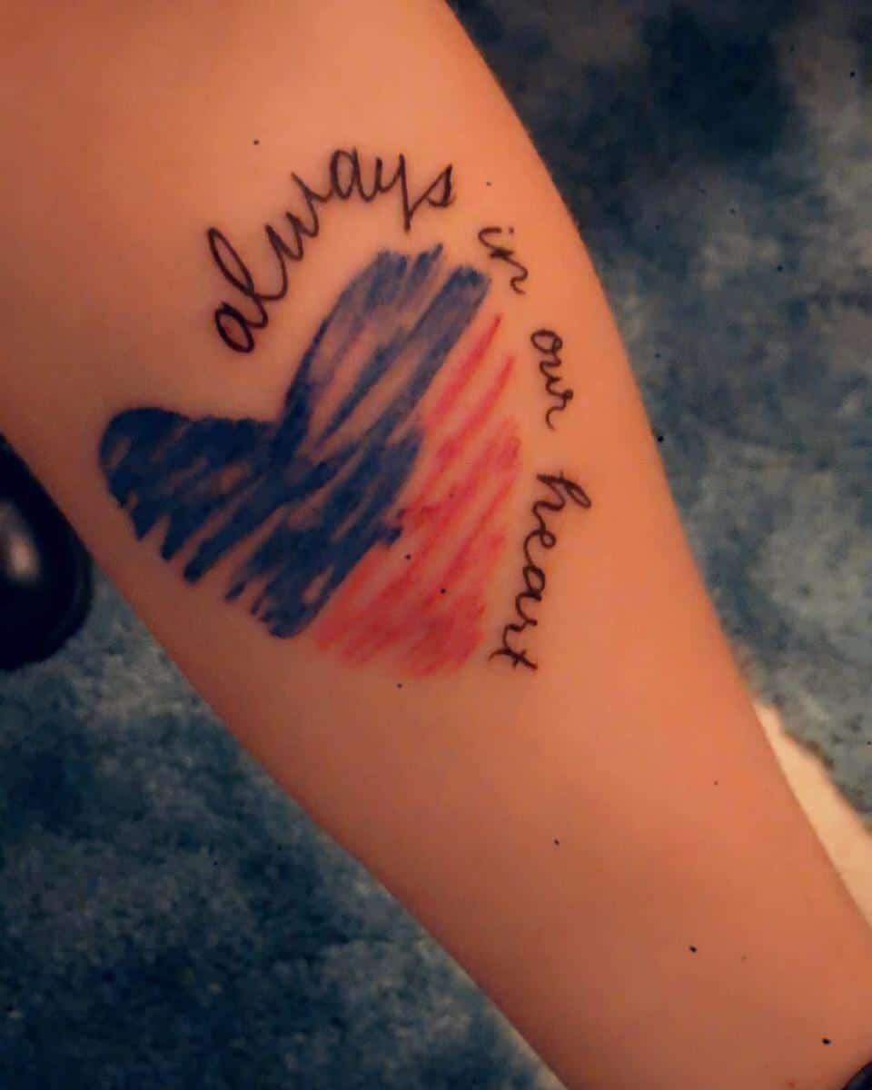 Tattoo of a heart with blue and red stripes and the words "always in our heart" on someone's arm