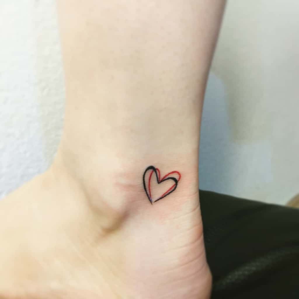 A minimalistic black and red heart tattoo on the inner side of an ankle