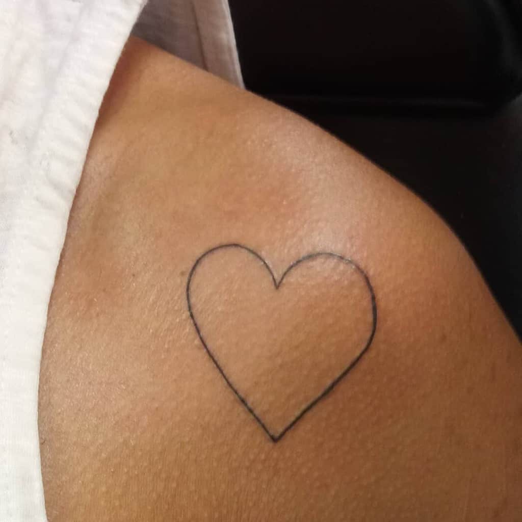 The tattoo features a clean and elegant black outline of a heart, placed just below the curve of the shoulder