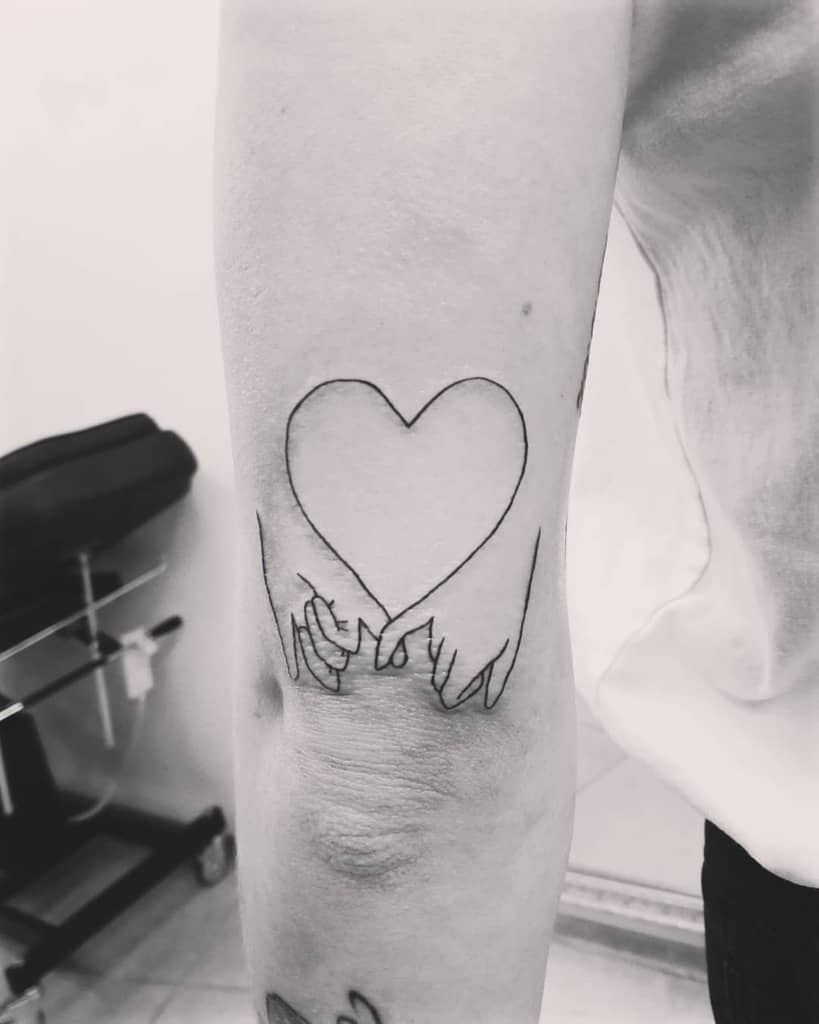 Black outline tattoo of hands holding pinkies forming a heart shape on an inner forearm
