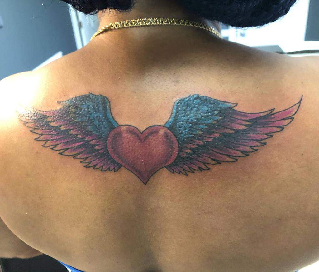 Heart with wings tattoo in shades of blue, pink, and red adorns the person's upper back