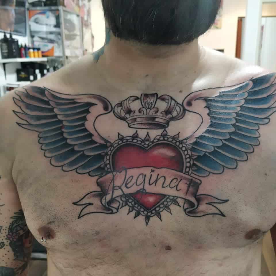 A tattoo with elegant wings, a heart, and a crown, all embraced by a banner reading "Regina" on a man's chest