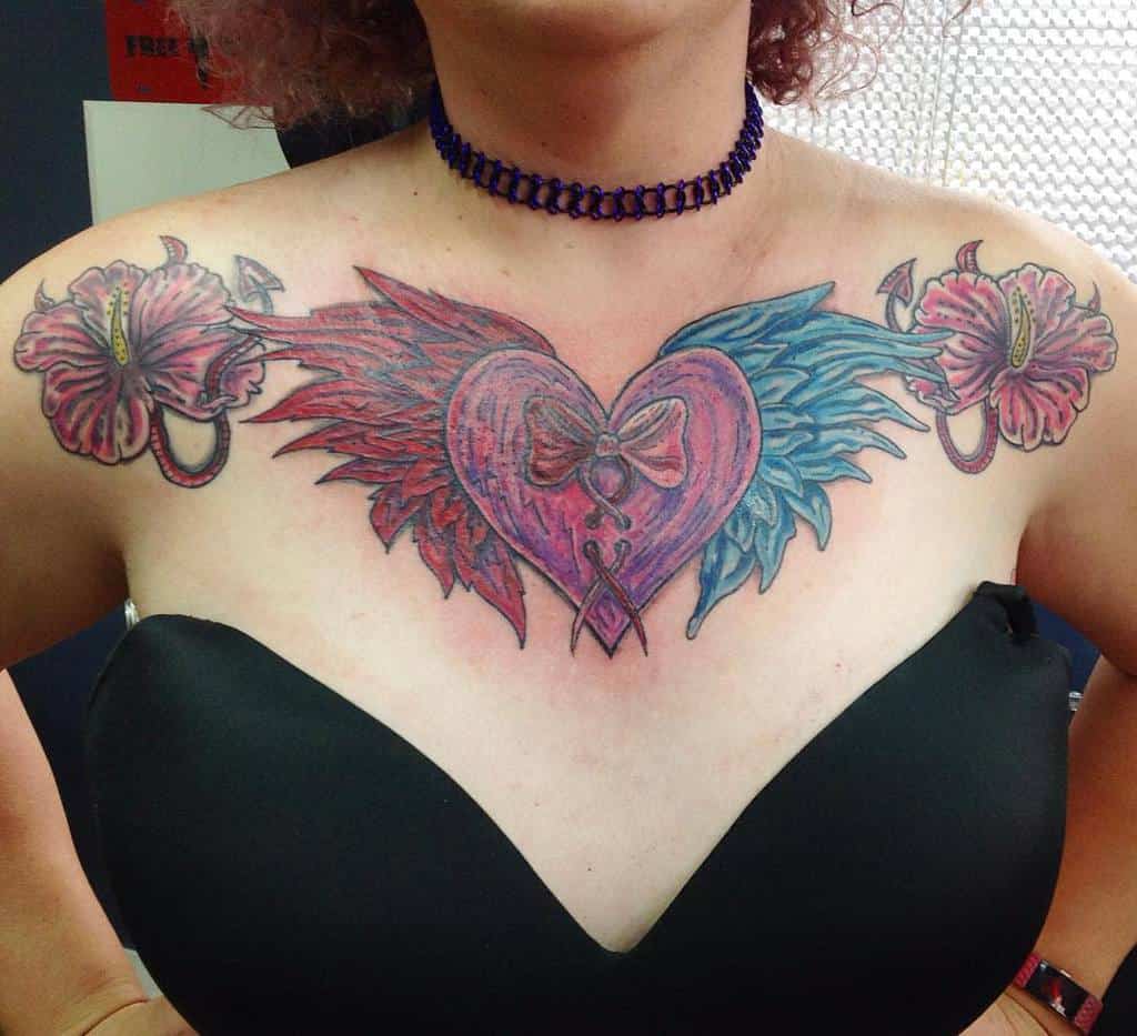 A colorful chest tattoo of a heart adorned with wings and flowers on a person wearing a black top
