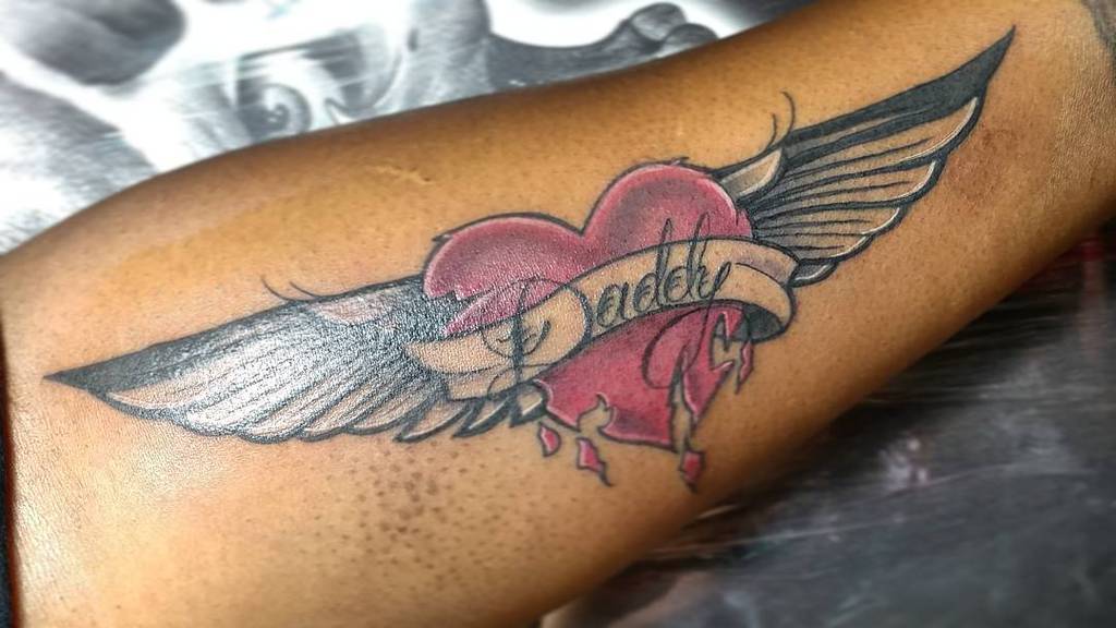 Tattoo of a winged heart with a banner reading "Daddy" on a person's arm
