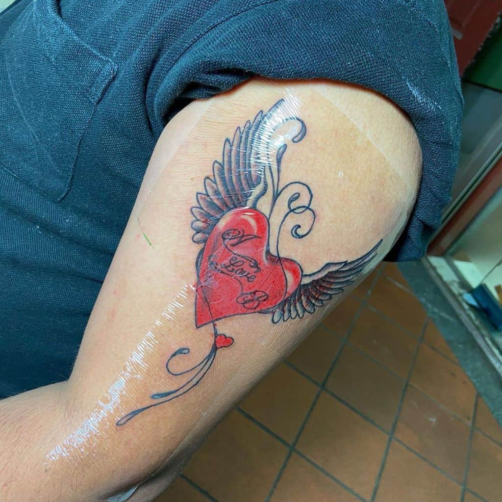 Tattoo of a heart with wings and text on a person's upper arm
