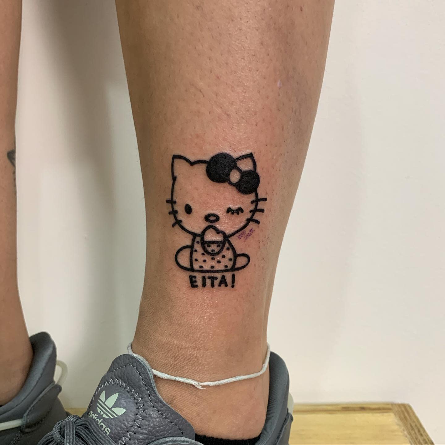 A tattoo of a winking Hello Kitty holding an ice cream cone with the word "Eita!" on a calf
