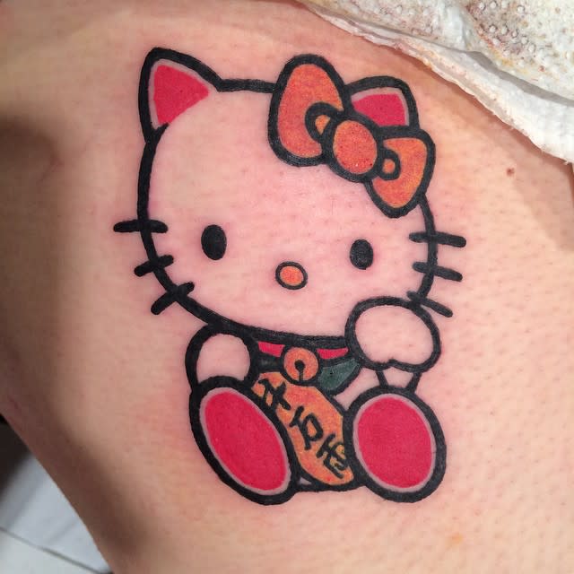Tattoo of Hello Kitty with a bow and a small gold charm featuring Japanese characters