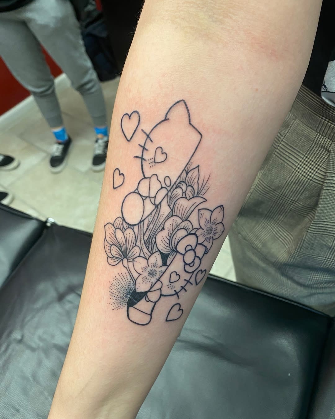 Outline tattoo of flowers and hearts with a Hello Kitty face on an inner forearm
