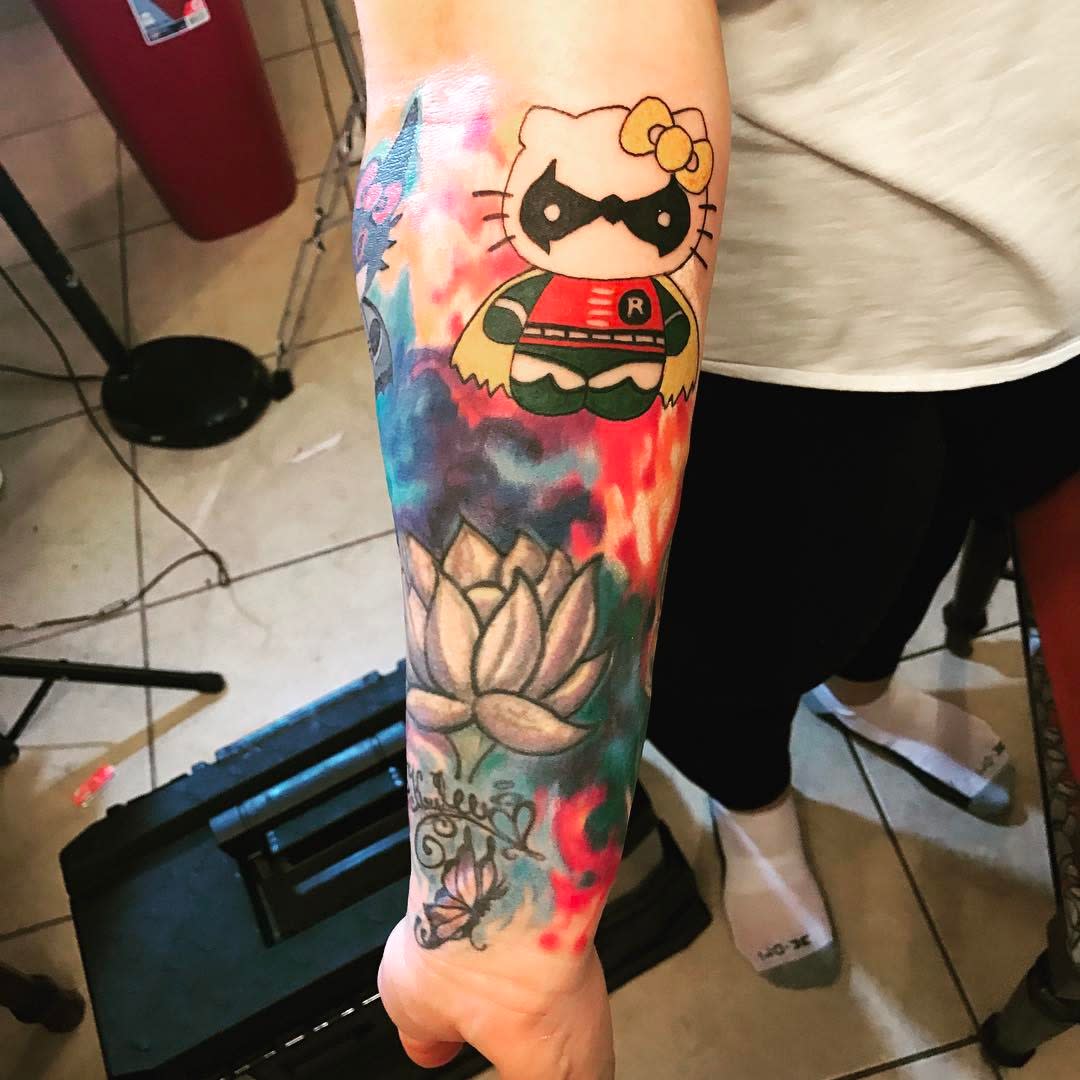 Tattoo of a Hello Kitty dressed as Robin on a forearm surrounded by colorful ink patterns