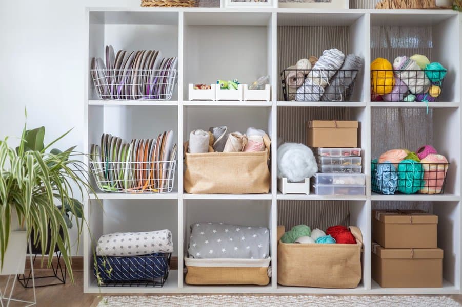 White cubby shelf with organized baskets, yarn, boxes, folded fabrics, and plates; plant on the left side