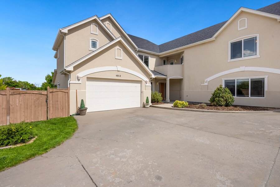 Large beige two-story house with white trim, attached garage, plants, and a wide driveway under a clear blue sky