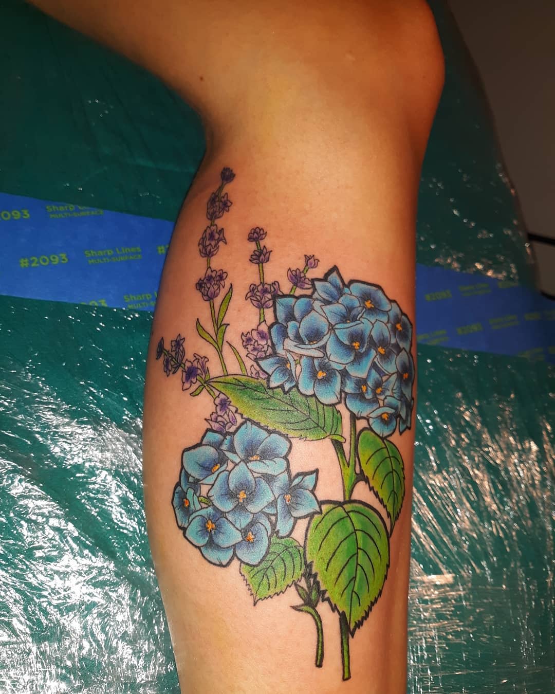 Tattoo of blue hydrangea flowers and purple accents on the back of a leg