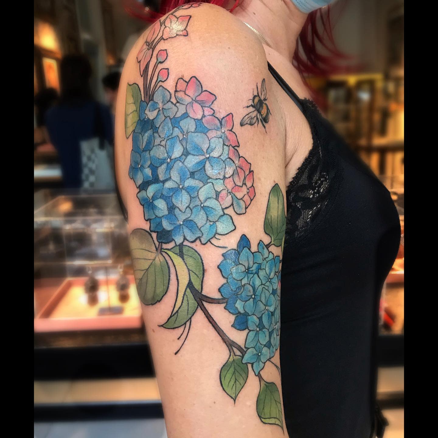 Upper arm tattoo of a blue hydrangea flower with a bee tattoo beside it
