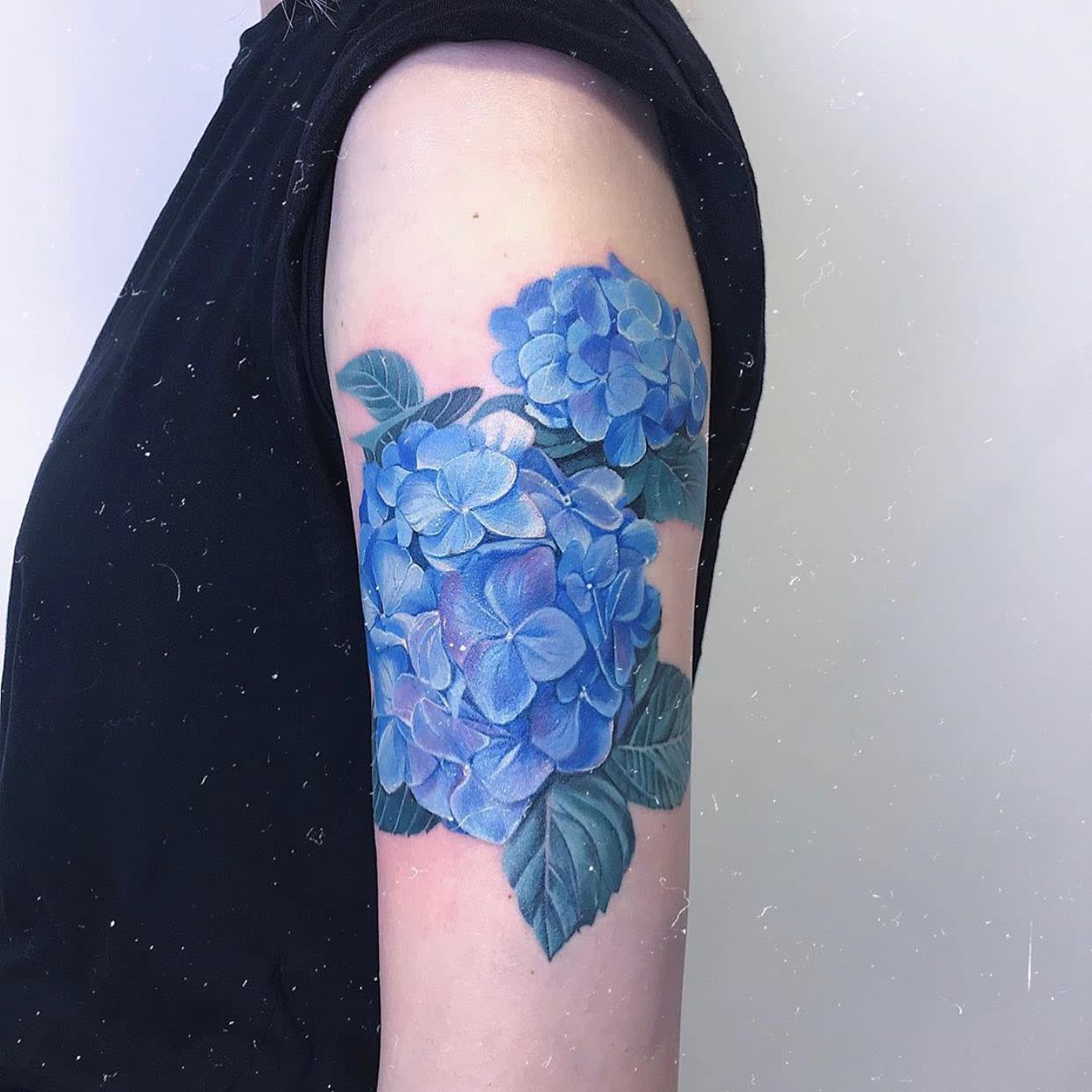 A detailed tattoo of blue hydrangeas on an upper arm, with green leaves visible