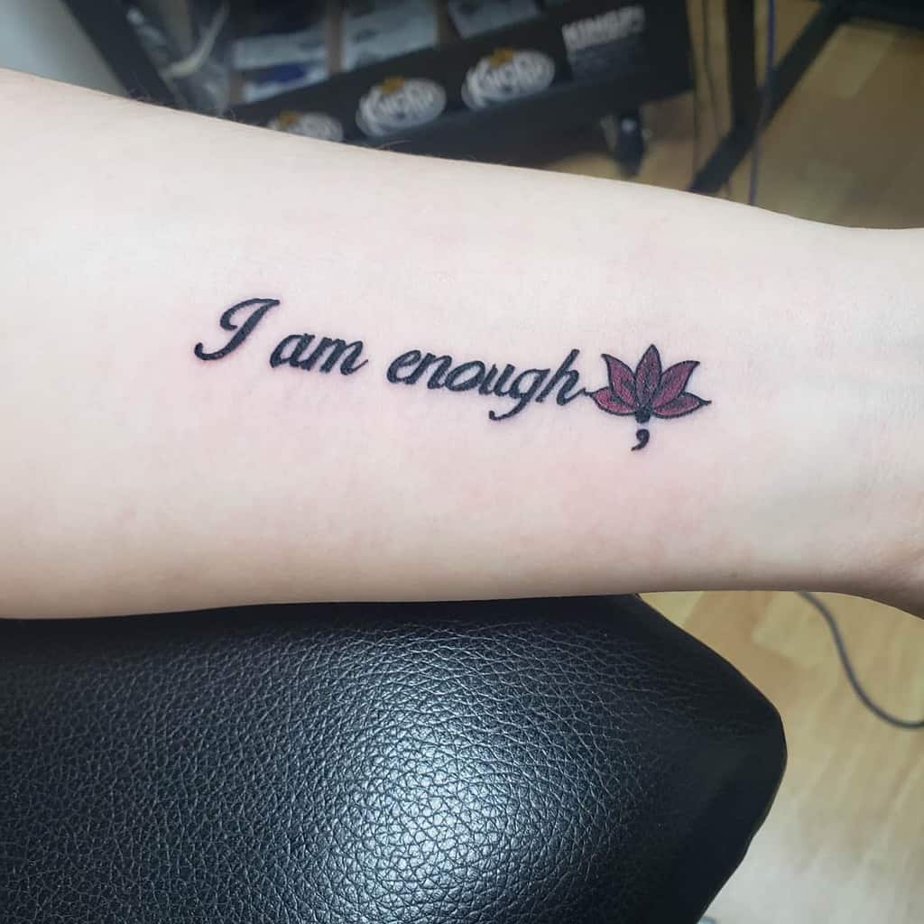 The tattoo features the empowering phrase "I am enough" with a delicate lotus flower and a semicolon at the end, symbolizing growth, strength, and self-worth.