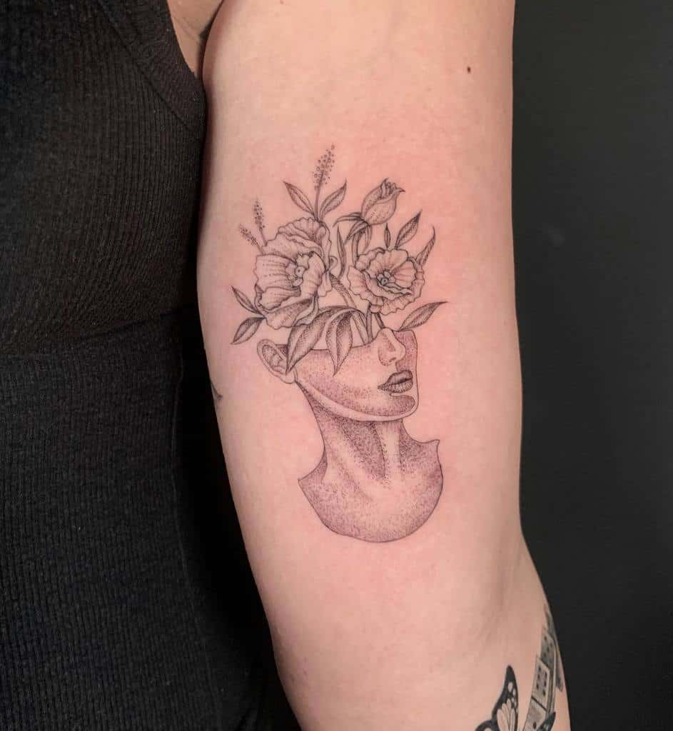 A single needle tattoo of a head with flowers blooming from the top, beautifully placed on a person's upper arm