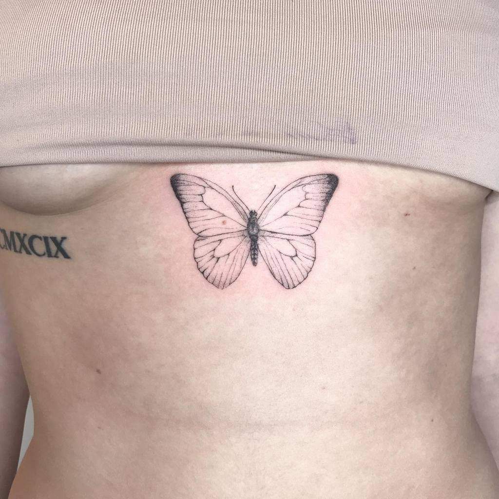 Delicate butterfly tattoo on the side of a person's ribcage artistically rendered with single needle precision accompanied by a Roman numeral tattoo beside it