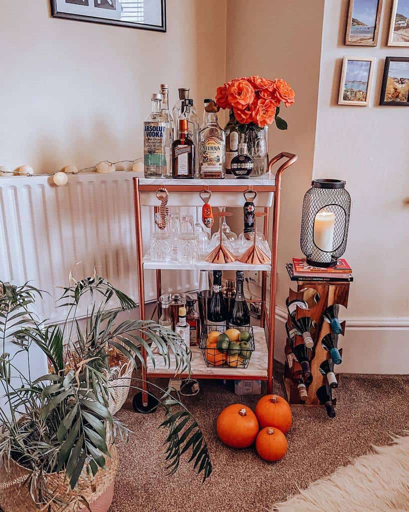 Industrial bar cart with liquor bottles, glasses, citrus fruits, and orange roses, next to pumpkins.