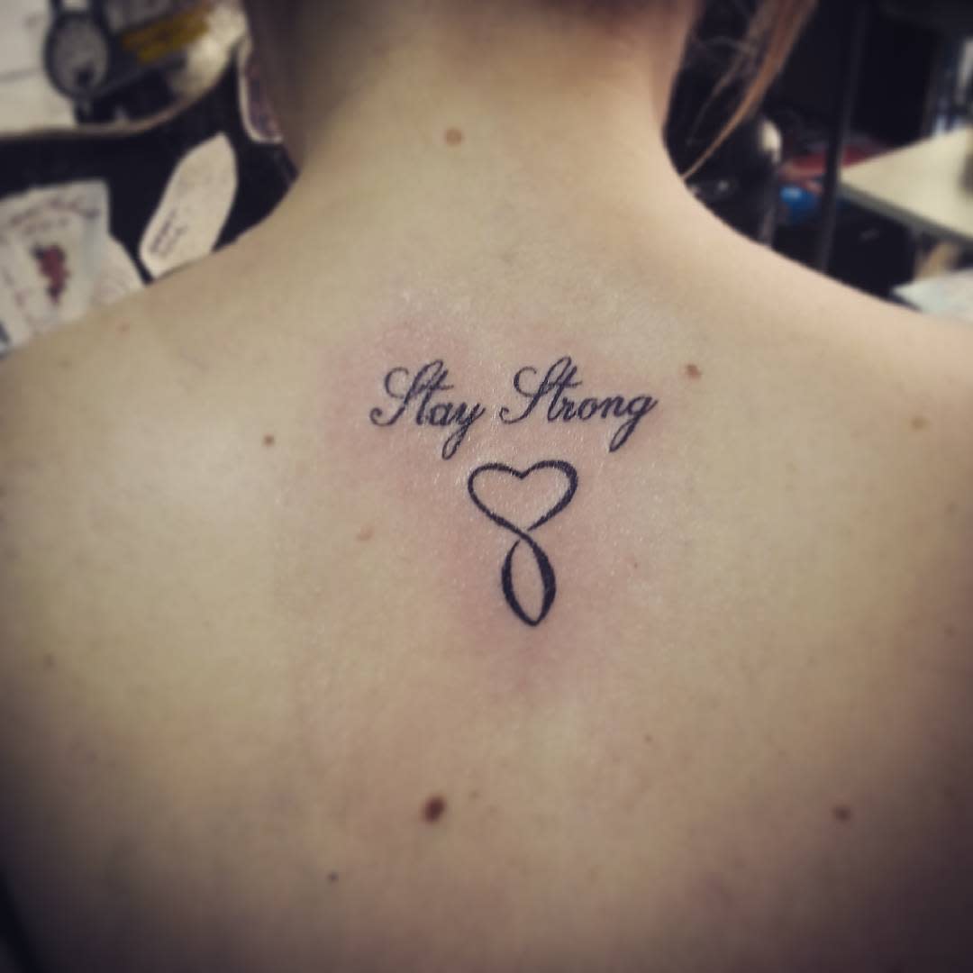 Tattoo on upper back reading "Stay Strong" with a heart-shaped ribbon below the text, subtly incorporating an infinity symbol