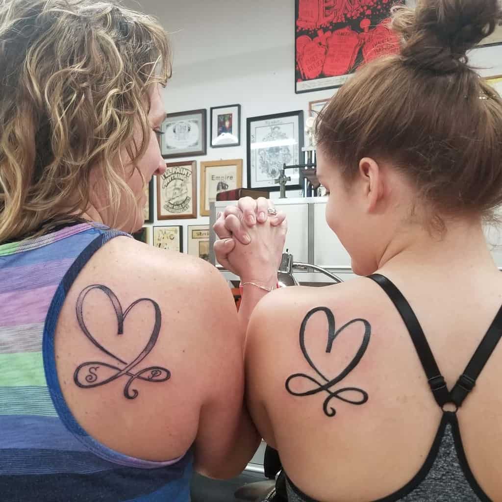 Two women proudly displaying matching heart tattoos on their shoulders, symbolizing an unbreakable bond as infinite as the love they share