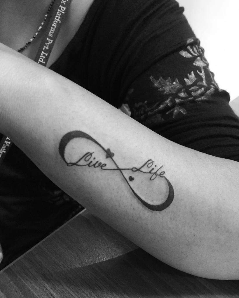 Black infinity tattoo with "Live Life" text and small hearts elegantly intertwined on a person's forearm