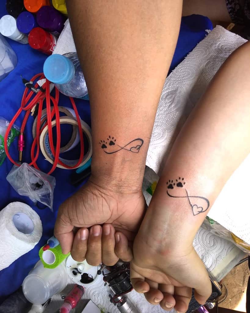 Two forearms with matching tattoos featuring an infinity symbol intertwined with paw prints and a heart