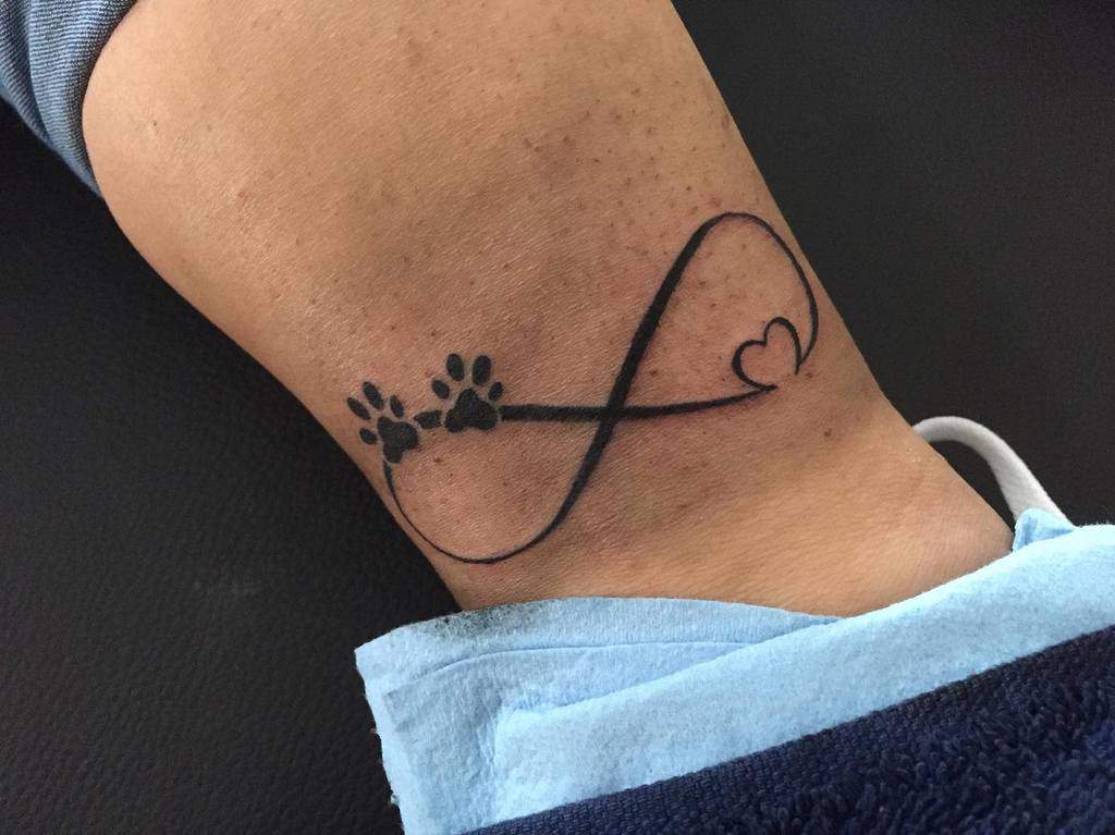 The tattoo on a lower leg showcases an infinity symbol intertwined with paw prints and a heart, integrated into the design