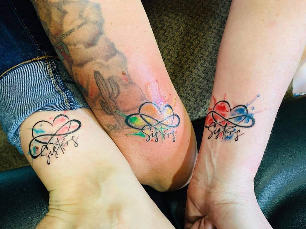 Three arms with matching "sisters" infinity heart tattoos in a vibrant watercolor style