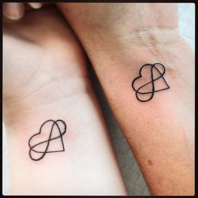 Two wrists with matching heart and infinity symbol tattoos in black ink