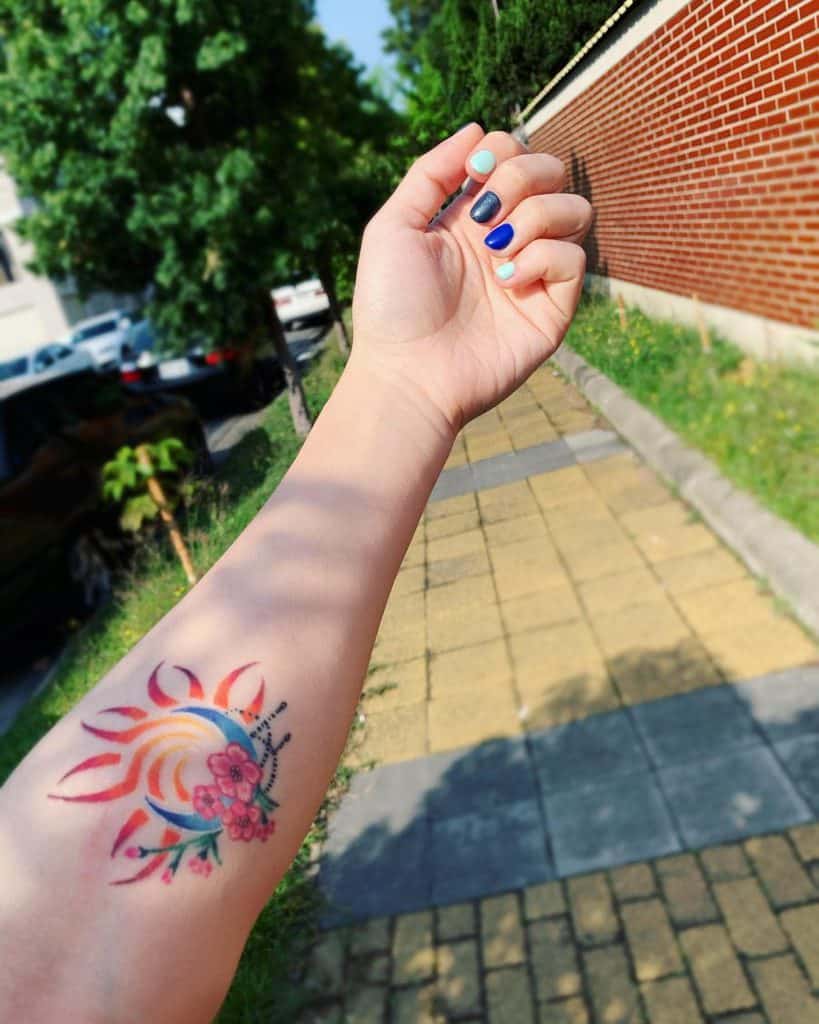 Colorful sun and moon tattoo on a forearm, featuring a crescent moon, fiery sun rays, and pink flowers with dotwork details.