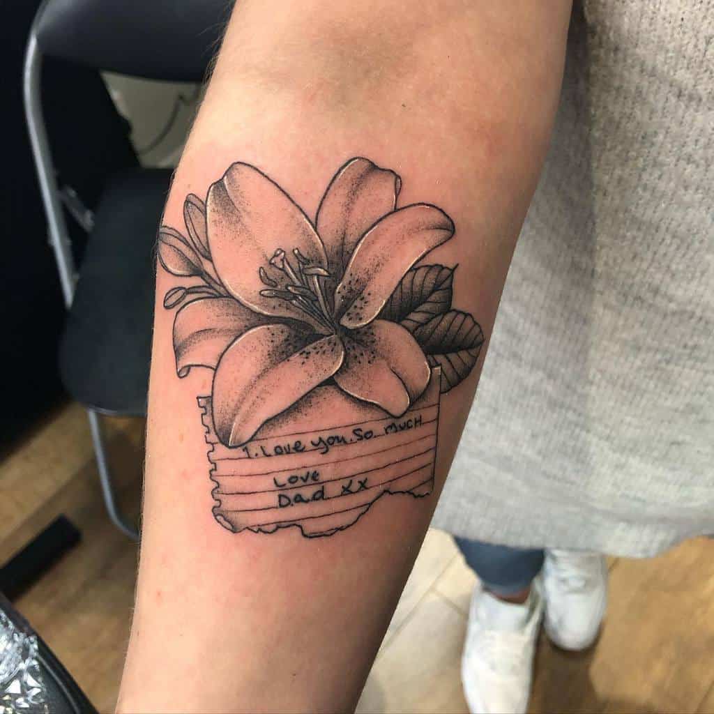 Heartfelt black and grey tattoo of a lily over a torn note with the handwritten message, 'I love you so much. Love, Dad xx'