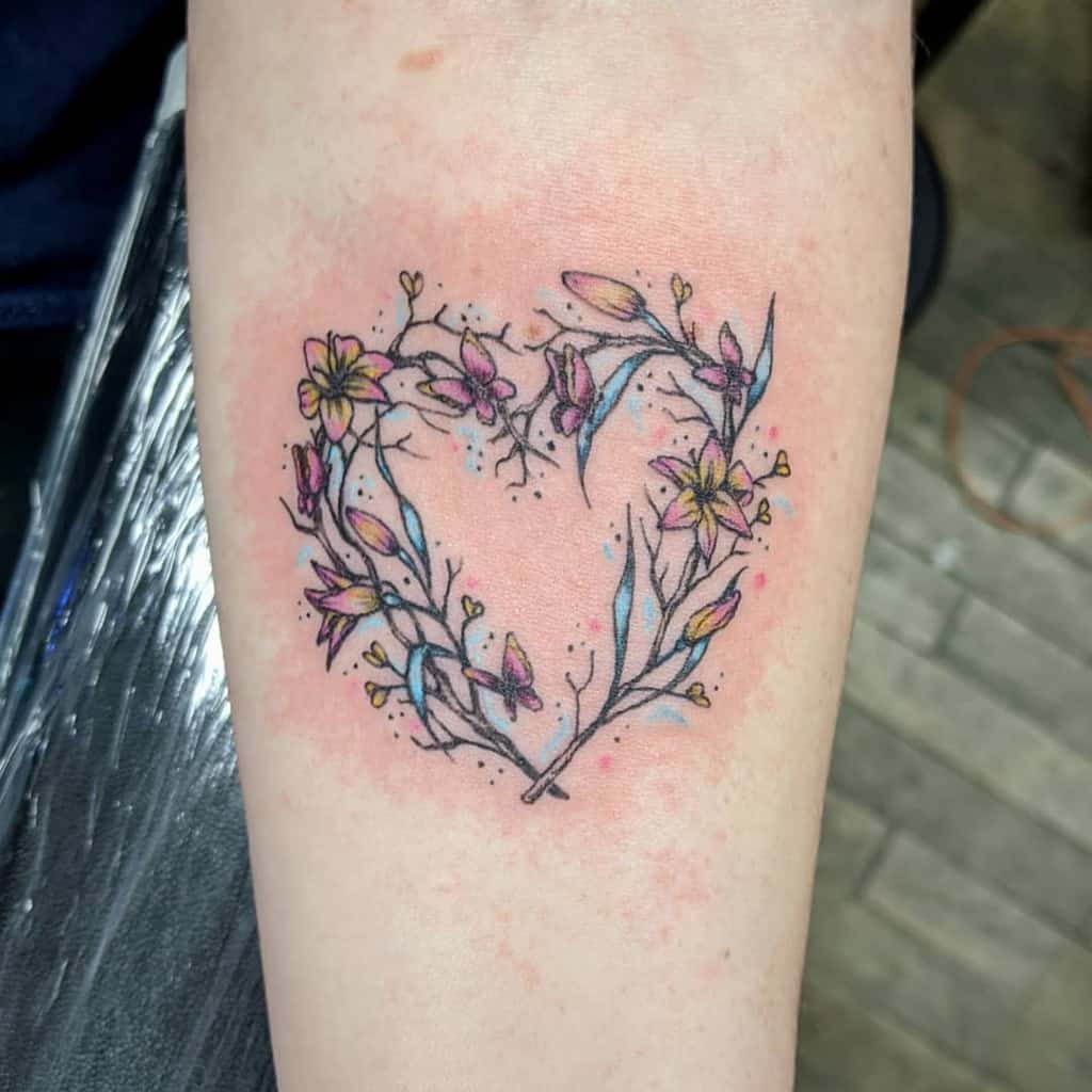 Delicate watercolor-style tattoo of a heart-shaped floral wreath of lilies with pink and yellow blossoms