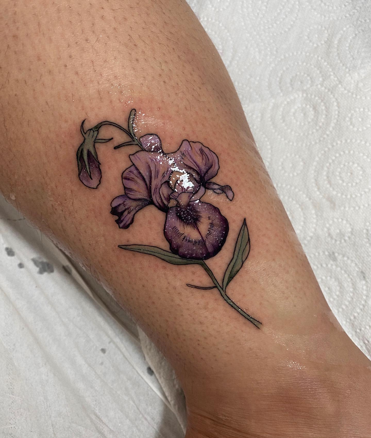 A purple iris tattoo design with green leaves on a leg