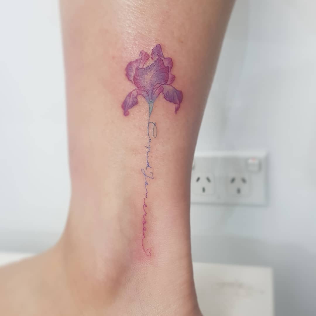 A delicate purple iris tattoo on an ankle with cursive text as the stem