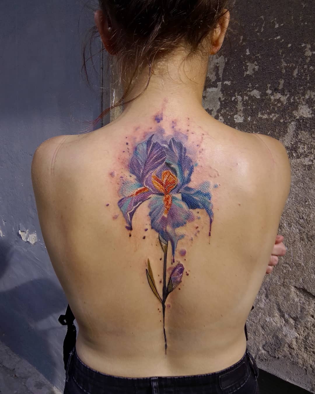 A vibrant tattoo of a large, colorful iris flower covers a woman's upper back and spine