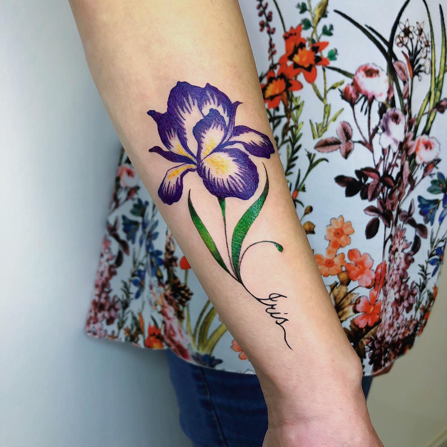 Forearm tattoo of a purple iris flower with green leaves and cursive text