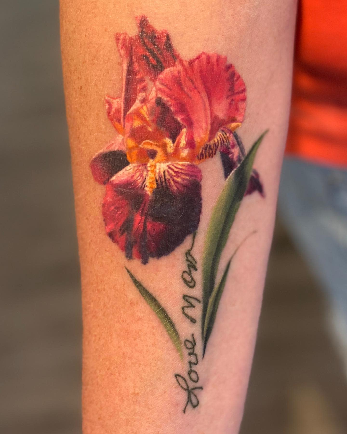 Tattoo of a colorful iris flower with "Love Mom" script on a forearm