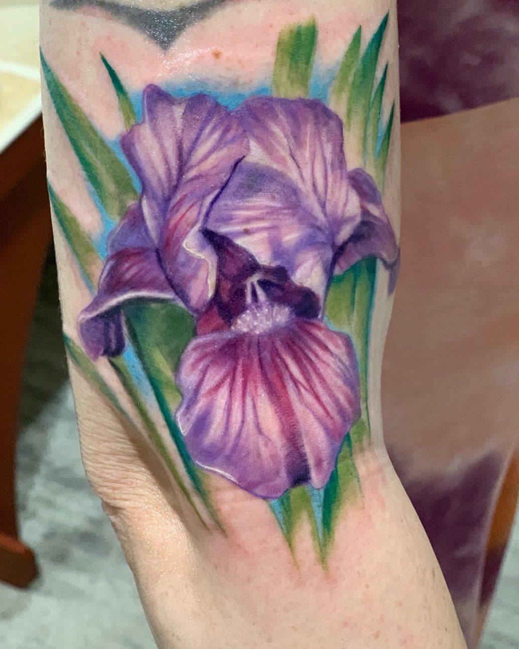 A tattoo of a purple iris flower with green leaves on a forearm