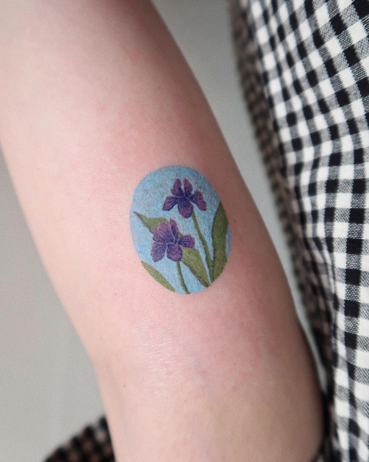 Round tattoo of purple iris flowers with green leaves on an arm