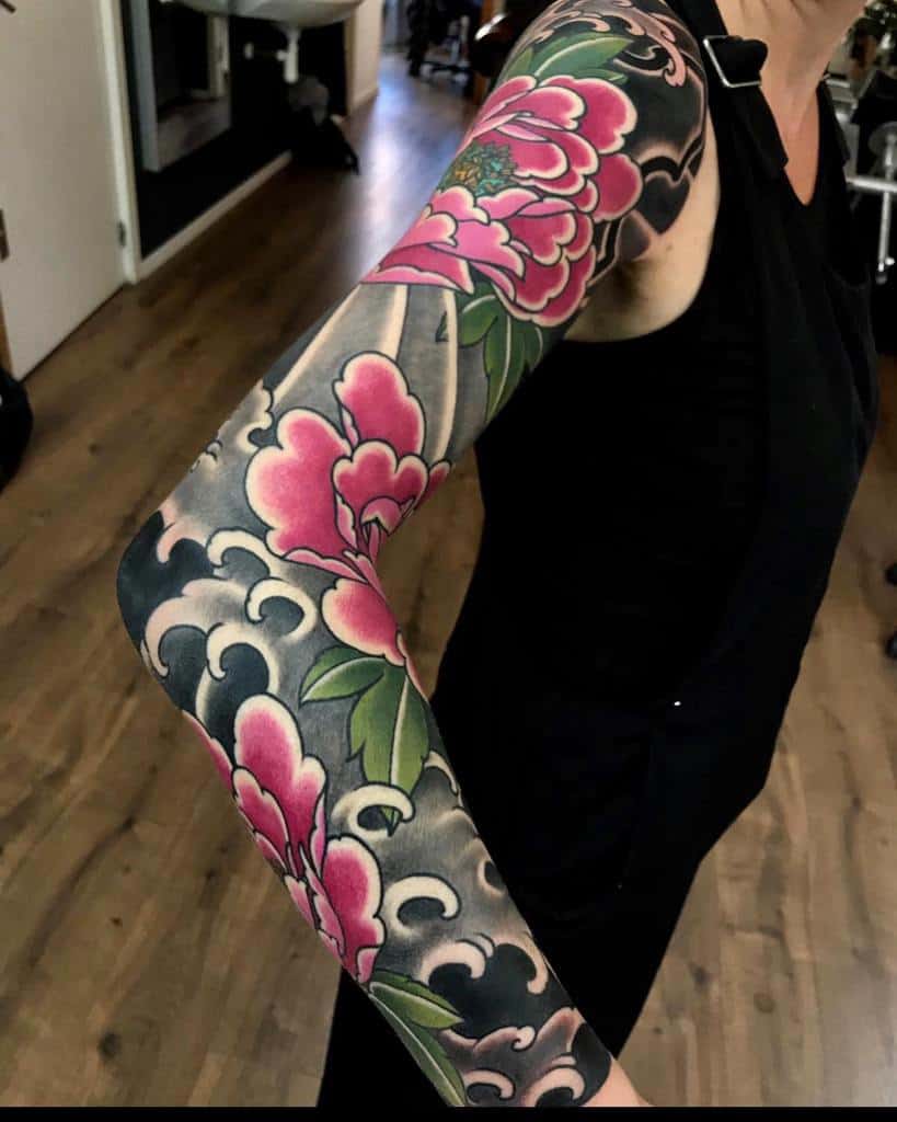 Japanese-style full sleeve tattoo featuring bold pink flowers, lush green leaves, and traditional cloud patterns with a dramatic black background for contrast