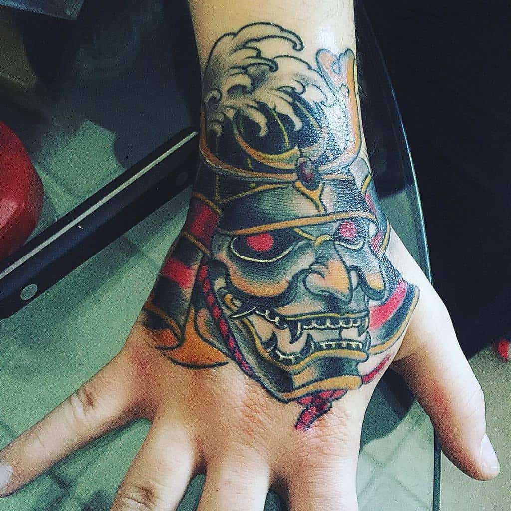 A hand tattoo of a ronin samurai mask in black, red, and gold covers the back of the hand