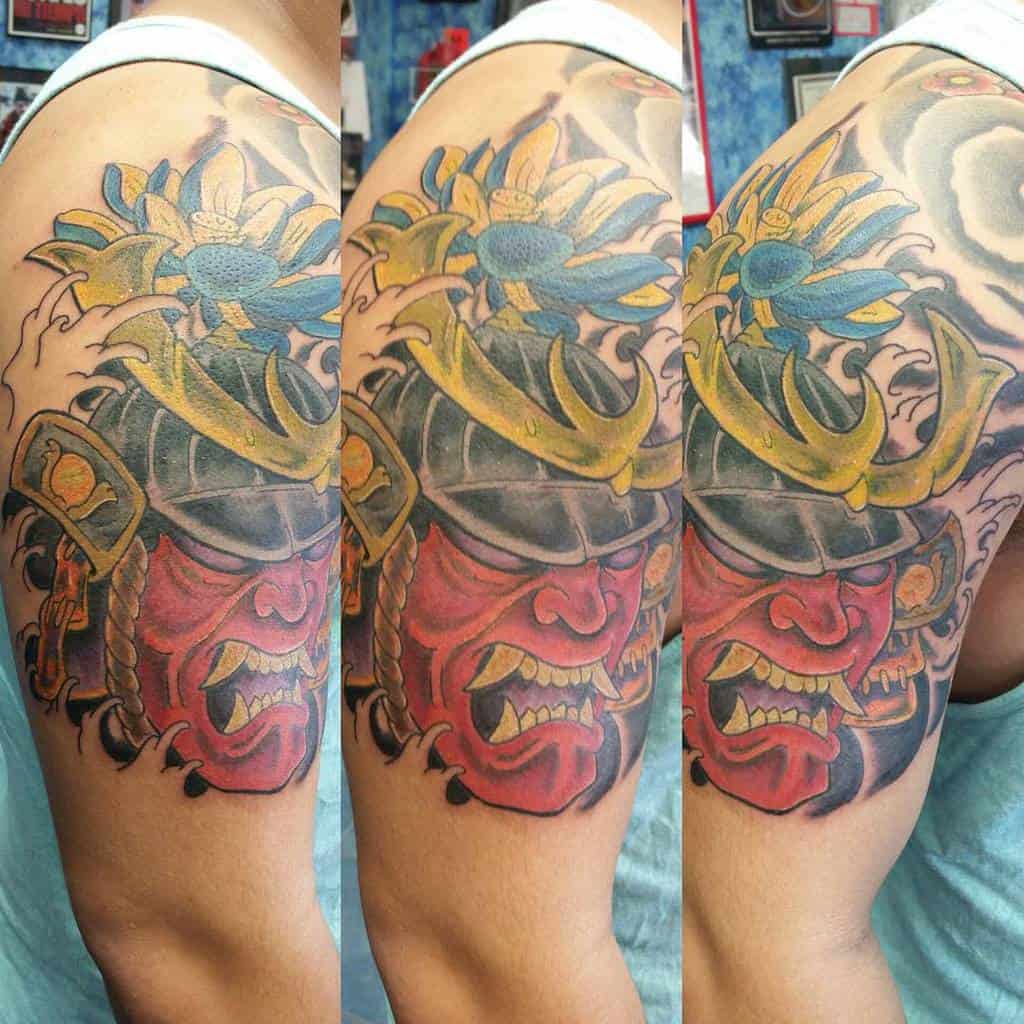 Colorful upper arm tattoo featuring a fierce red samurai mask adorned with golden armor details and a vibrant blue lotus flower