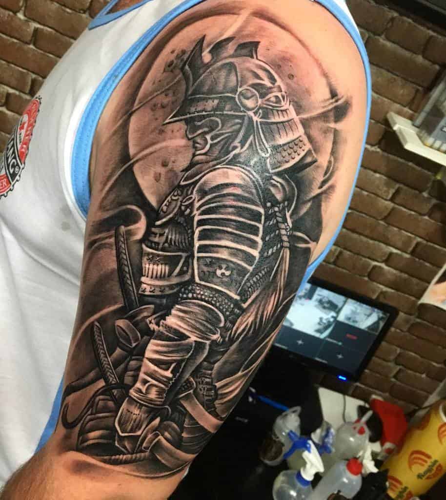 Detailed black and grey upper arm tattoo depicting a samurai in traditional armor, with intricate shading and highlights, giving a dynamic, three-dimensional effect