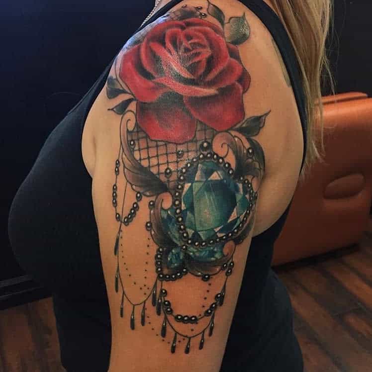Ornate tattoo featuring a realistic red rose, a large blue gemstone, black lace details, and chandelier-style beadwork for an elegant design
