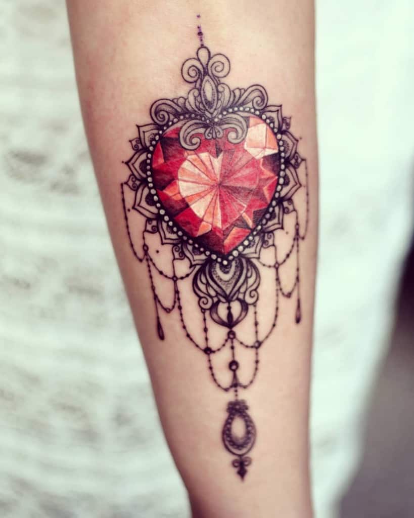 Elegant tattoo featuring a faceted red gemstone heart encased in ornate filigree, chandelier-style beads, and intricate decorative accents