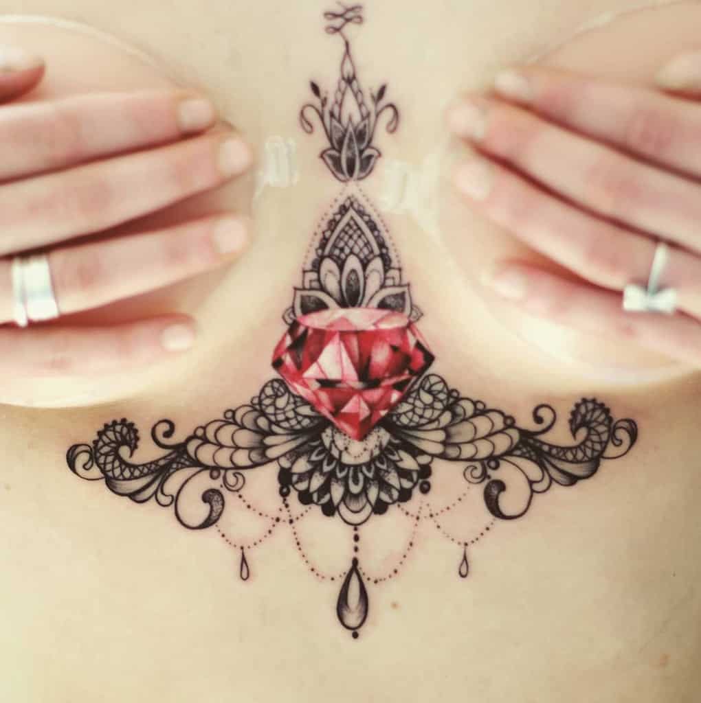 A tattoo featuring a faceted red diamond gemstone framed by intricate filigree, mandala details, and chandelier-style teardrop beads