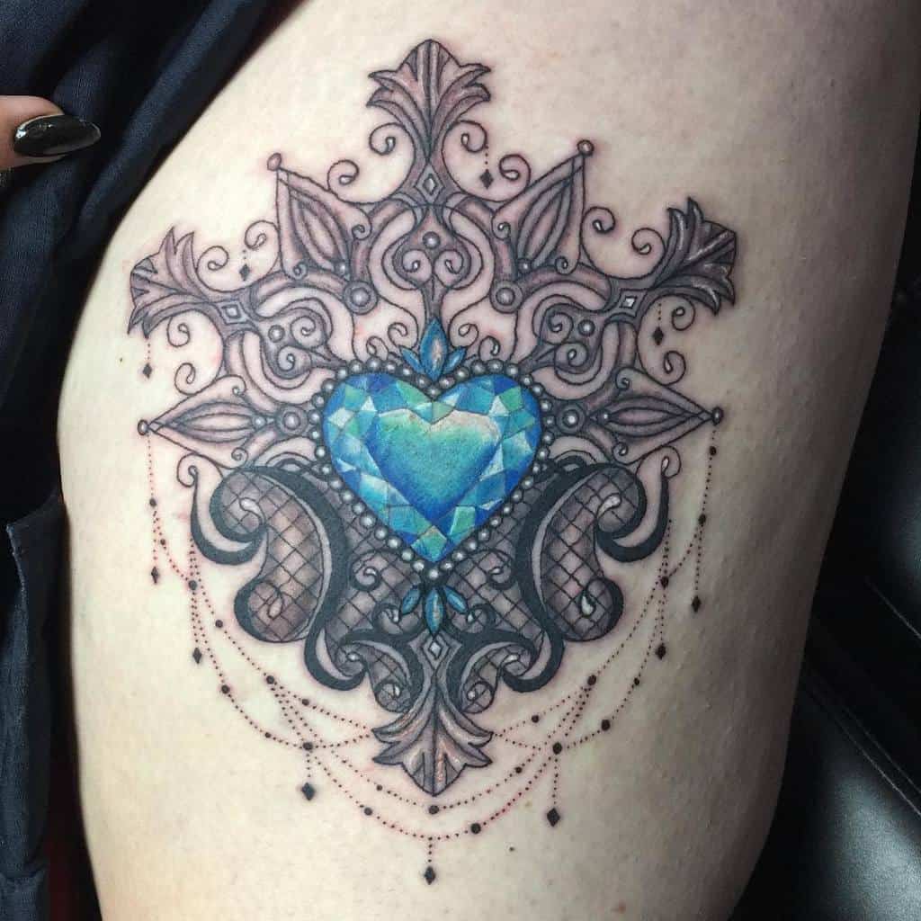 A tattoo featuring a faceted blue heart-shaped diamond gemstone framed by intricate baroque-style filigree, pearls, and chandelier-style teardrop beads