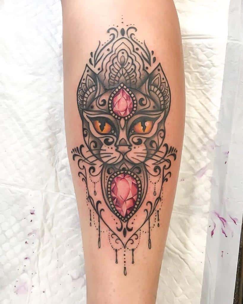 Ornate tattoo of a cat's face with piercing orange eyes, intricate filigree details, two faceted pink gemstones, and chandelier-style teardrop beads