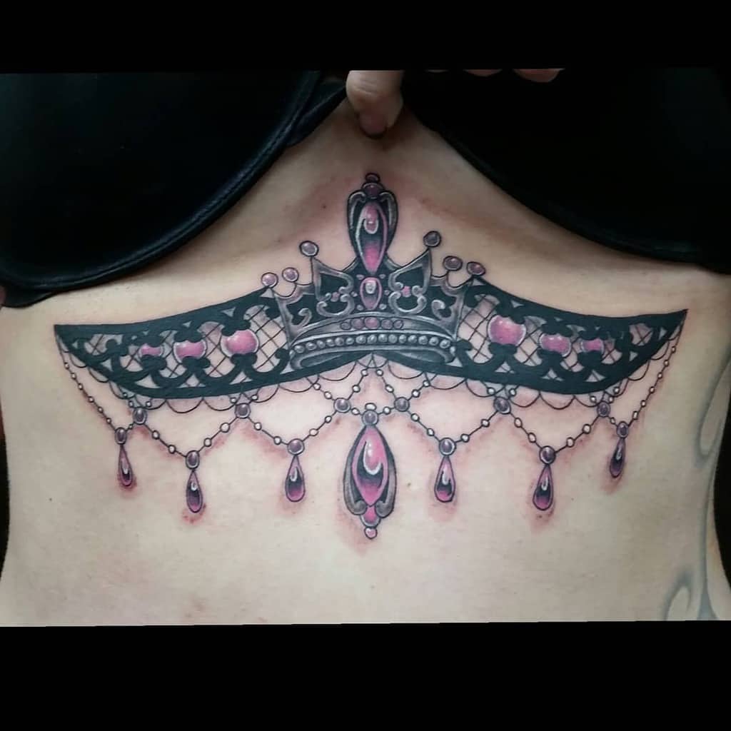 A tattoo featuring a black lace design with a jeweled crown at the center, pink gemstone accents, and chandelier-style teardrop beads