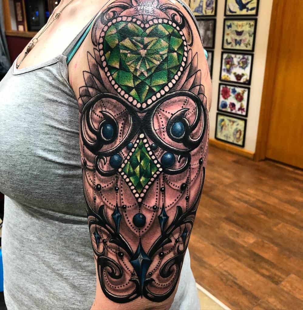 Intricate tattoo designed to resemble an owl, featuring a large faceted green heart-shaped gemstone as its centerpiece, ornate filigree, blue jewel accents, and chandelier-style beadwork